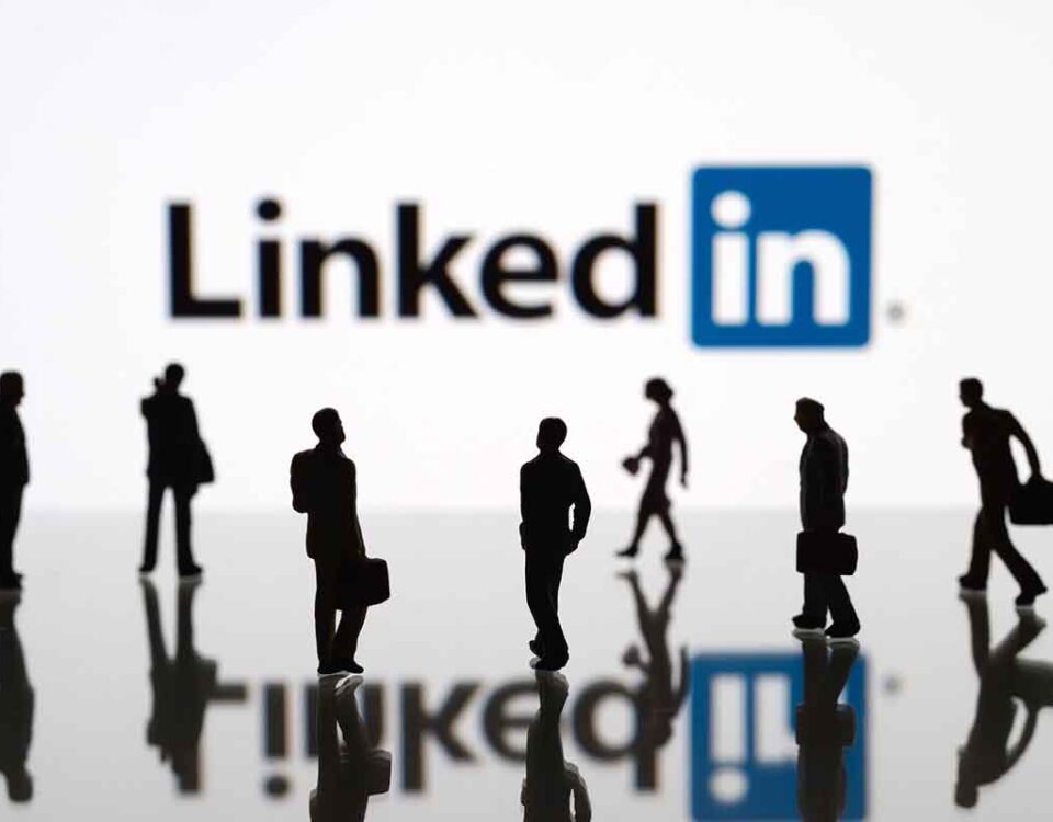 Pessoas com roupas de trabalho, de executivos, andando, e no fundo o logo do LinkedIn