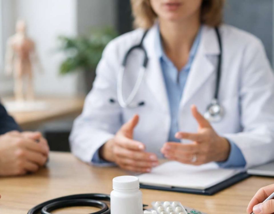 Consultório médico com um profissional discutindo com pacientes. Medicamentos e computador na mesa.